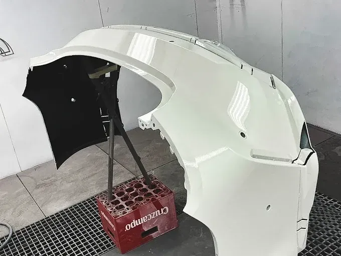 Un parachoques de coche blanco brillante montado sobre un soporte dentro de una cabina de pintura.
