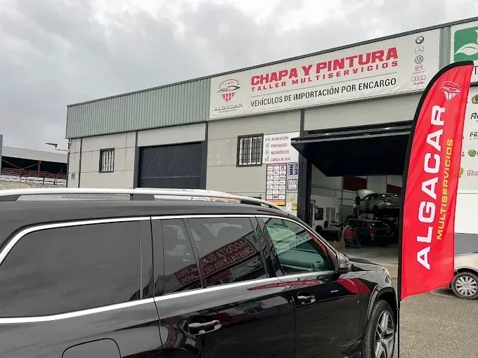 Un SUV negro estacionado frente al taller de carrocería Algacar con una bandera promocional roja a la derecha.