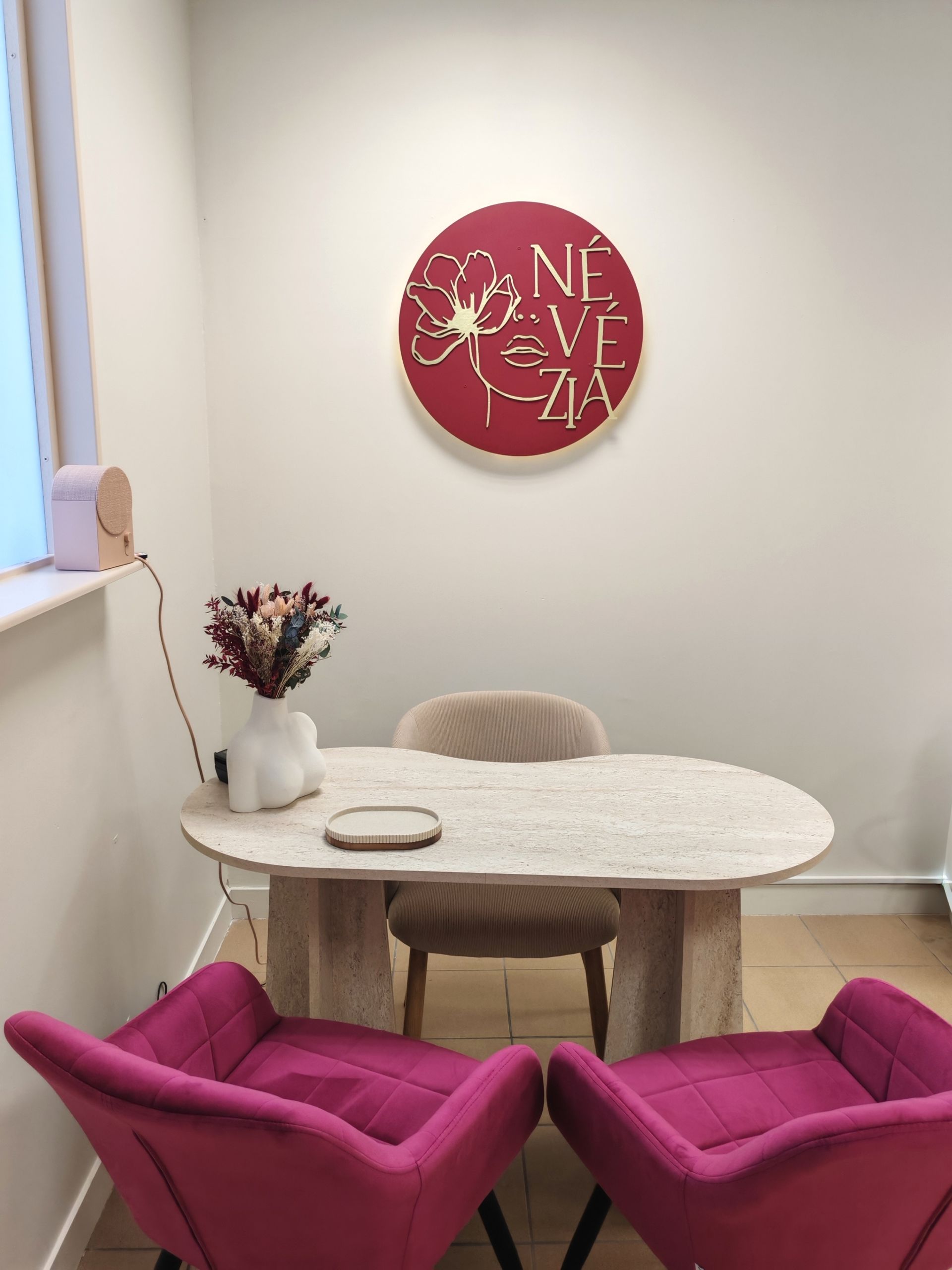 Petit coin réunion avec chaises rouges et roses, table ovale, décorations murales et fleurs près d'une fenêtre.