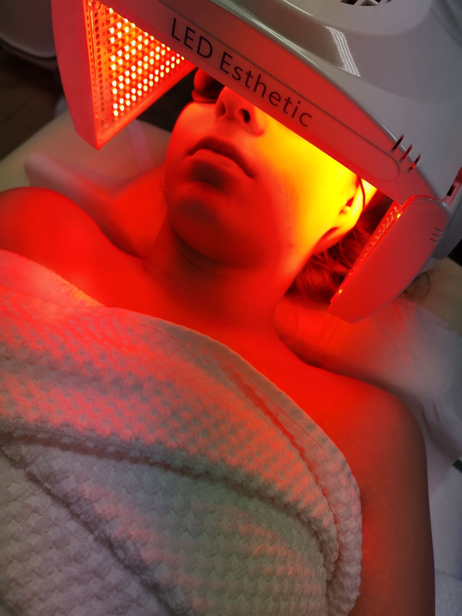 Personne allongée sous un appareil de traitement par masque facial à LED rouges, enveloppée dans une serviette.