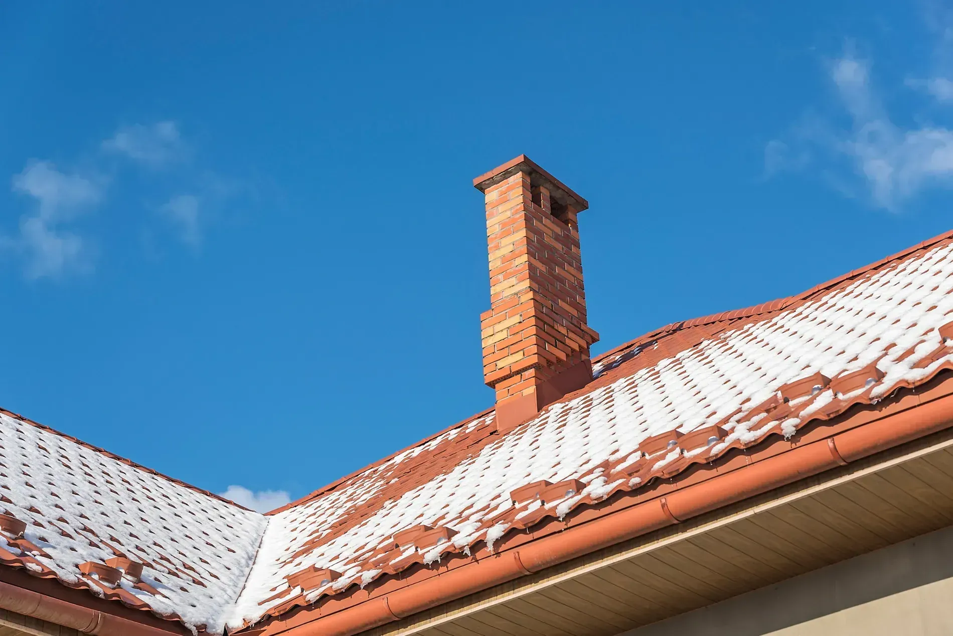 Una chimenea de ladrillo que se eleva desde un tejado de tejas rojas parcialmente cubierto de nieve ligera bajo un cielo azul despejado.