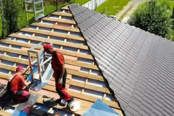 Dos trabajadores con uniformes rojos instalan tejas en el tejado inclinado de una vivienda con vigas de madera a la vista.