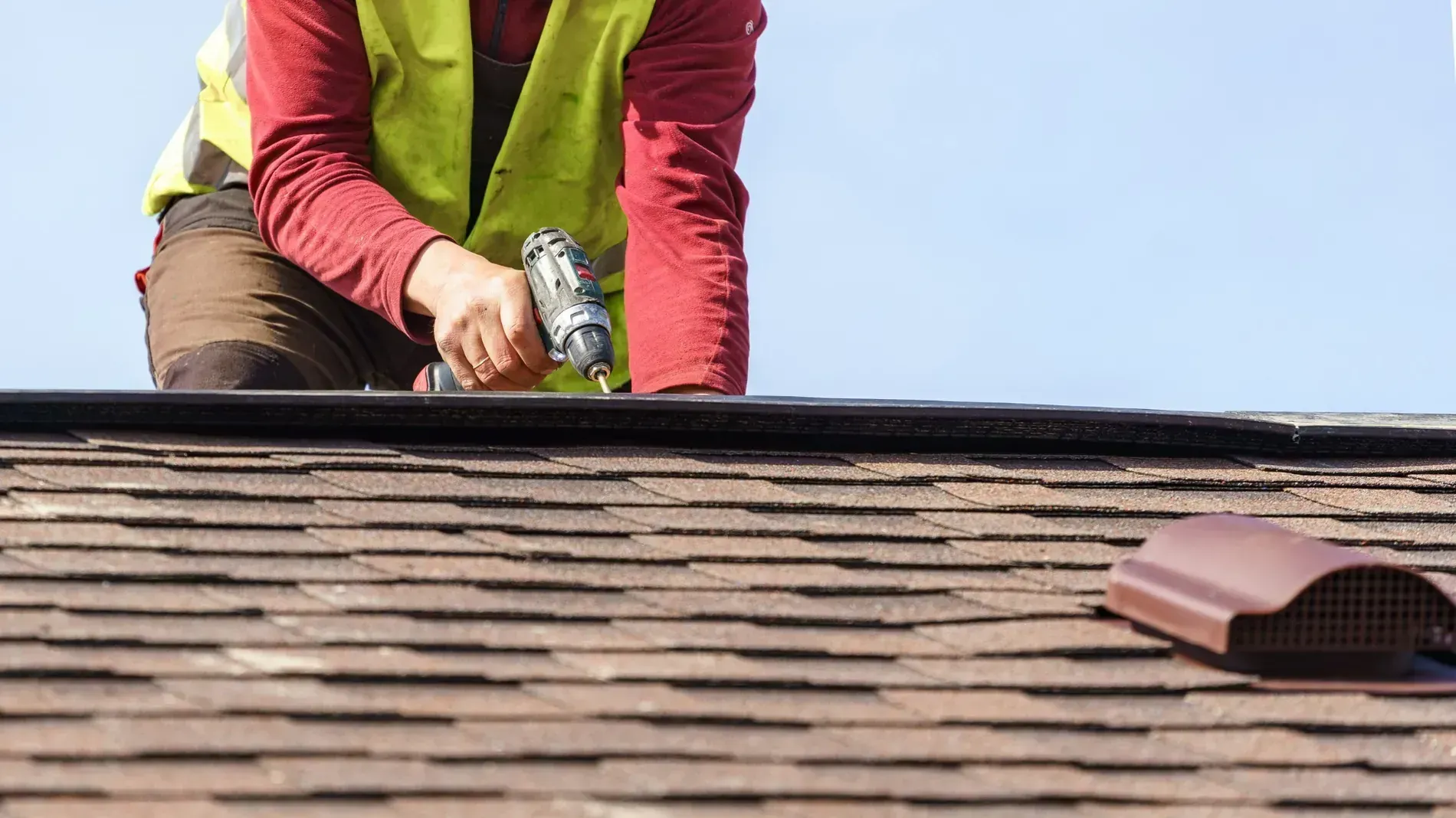 Un trabajador con un chaleco reflectante utiliza un taladro eléctrico para instalar tejas en un tejado residencial marrón.