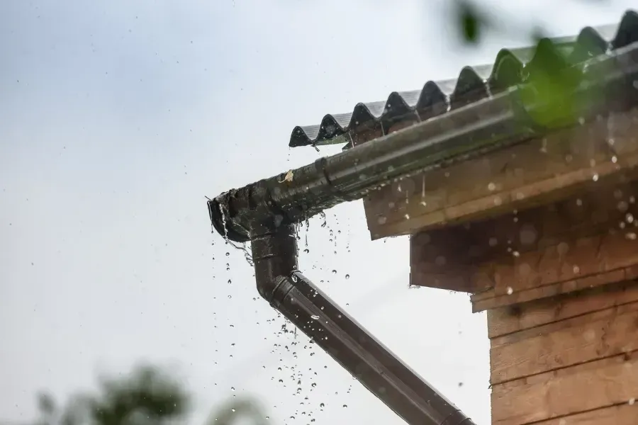 Agua de lluvia desbordándose de una canaleta oscura y obstruida adosada a un edificio de madera en un día lluvioso.