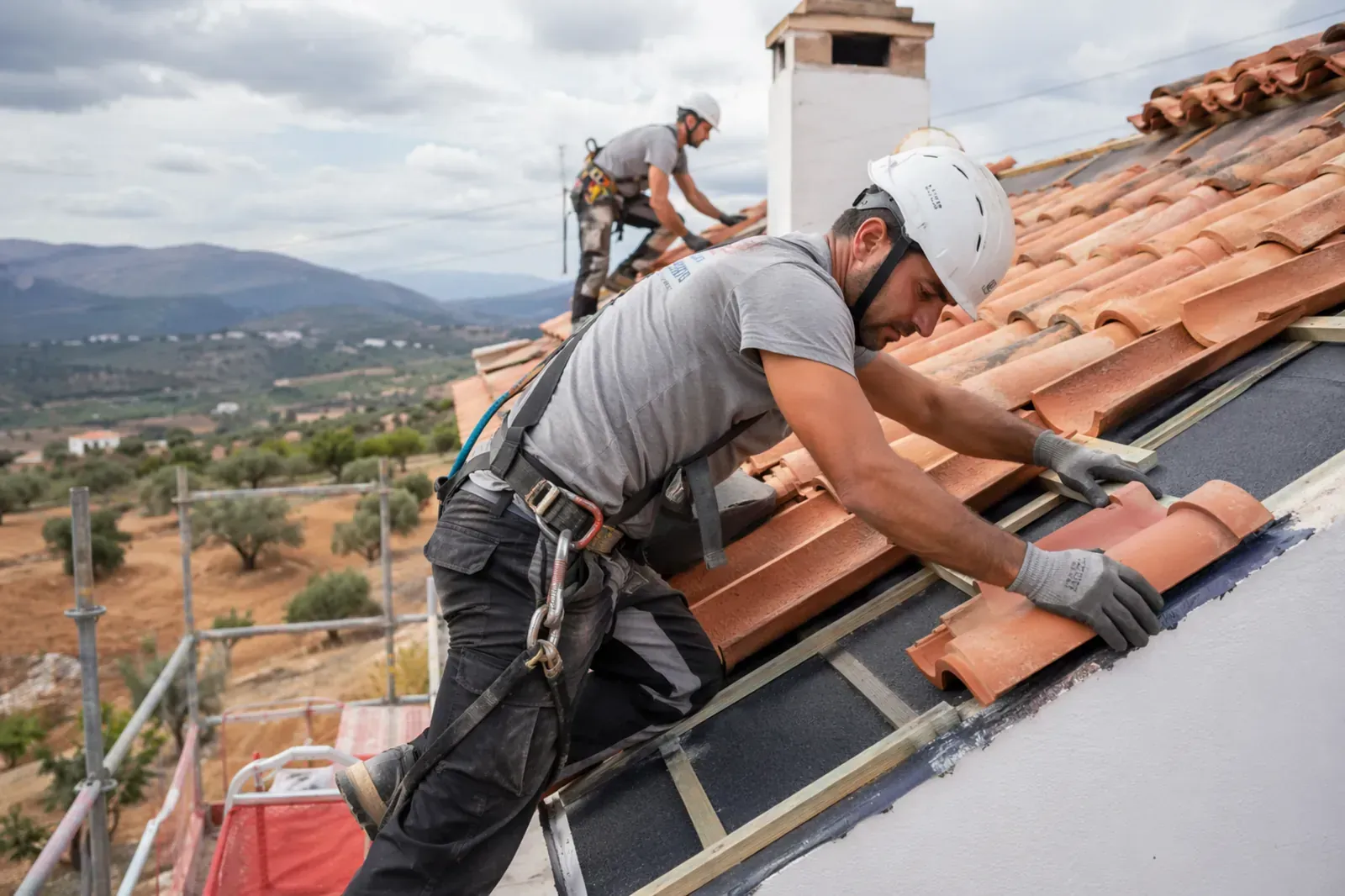 Dos obreros de la construcción, equipados con trajes de seguridad, instalan tejas de terracota en un tejado inclinado y soleado.
