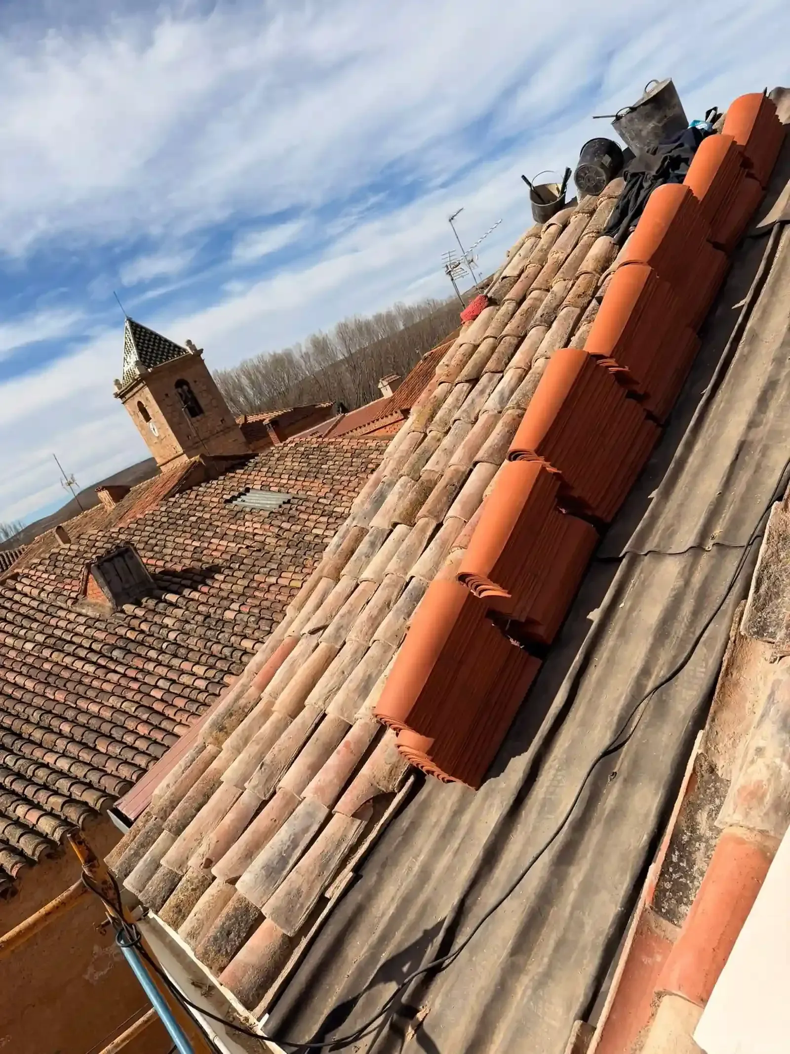 Vista desde la azotea de tejas de terracota desgastadas, con una hilera de tejas rojas nuevas colocadas y un campanario a lo lejos al fondo.