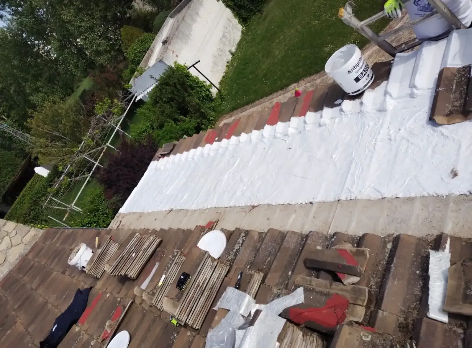 Una persona con guantes de trabajo aplica una membrana selladora impermeable blanca a un tejado de tejas en un día soleado.