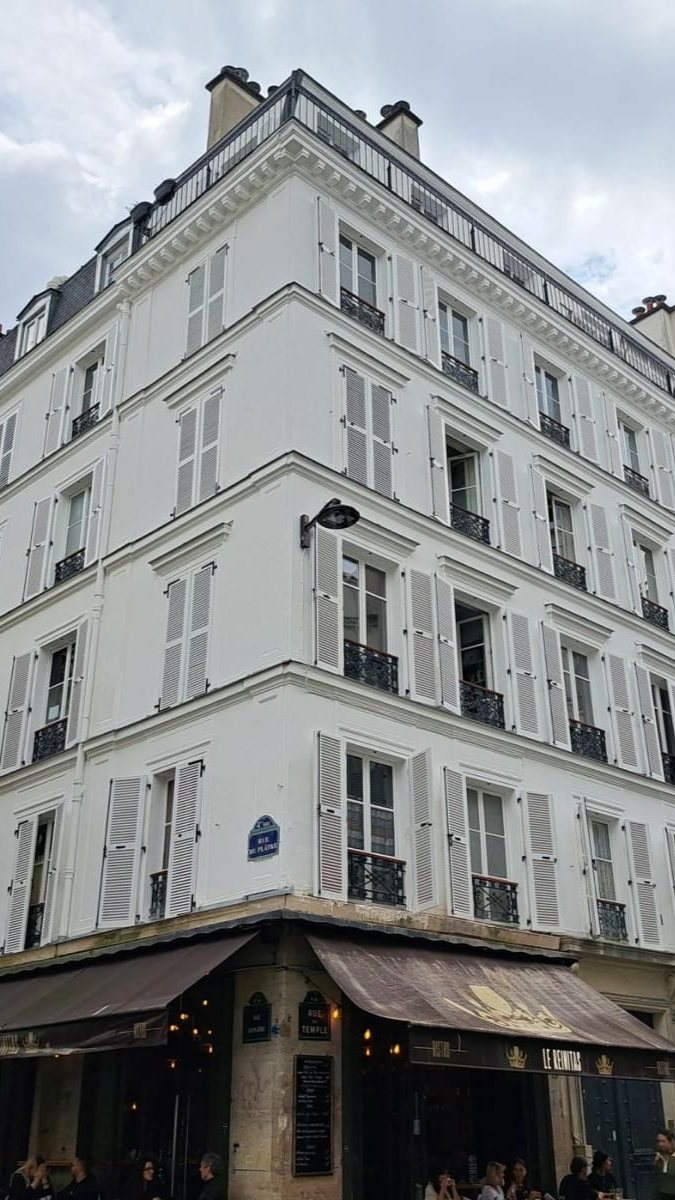 Immeuble parisien blanc à volets, café en dessous. Ciel nuageux.
