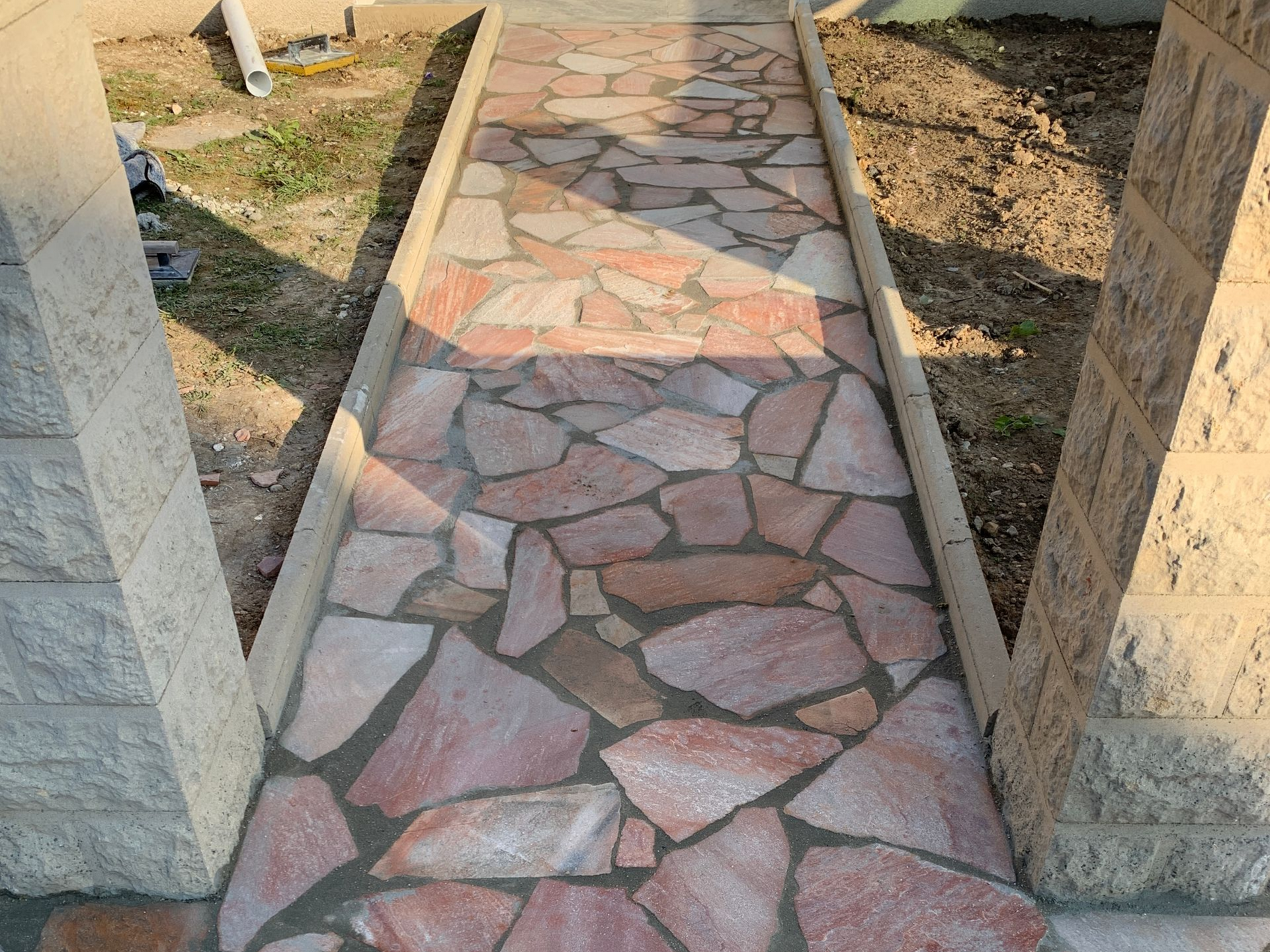 Allée en dalles de pierre rose et beige, encadrée de bordures et de piliers en béton.