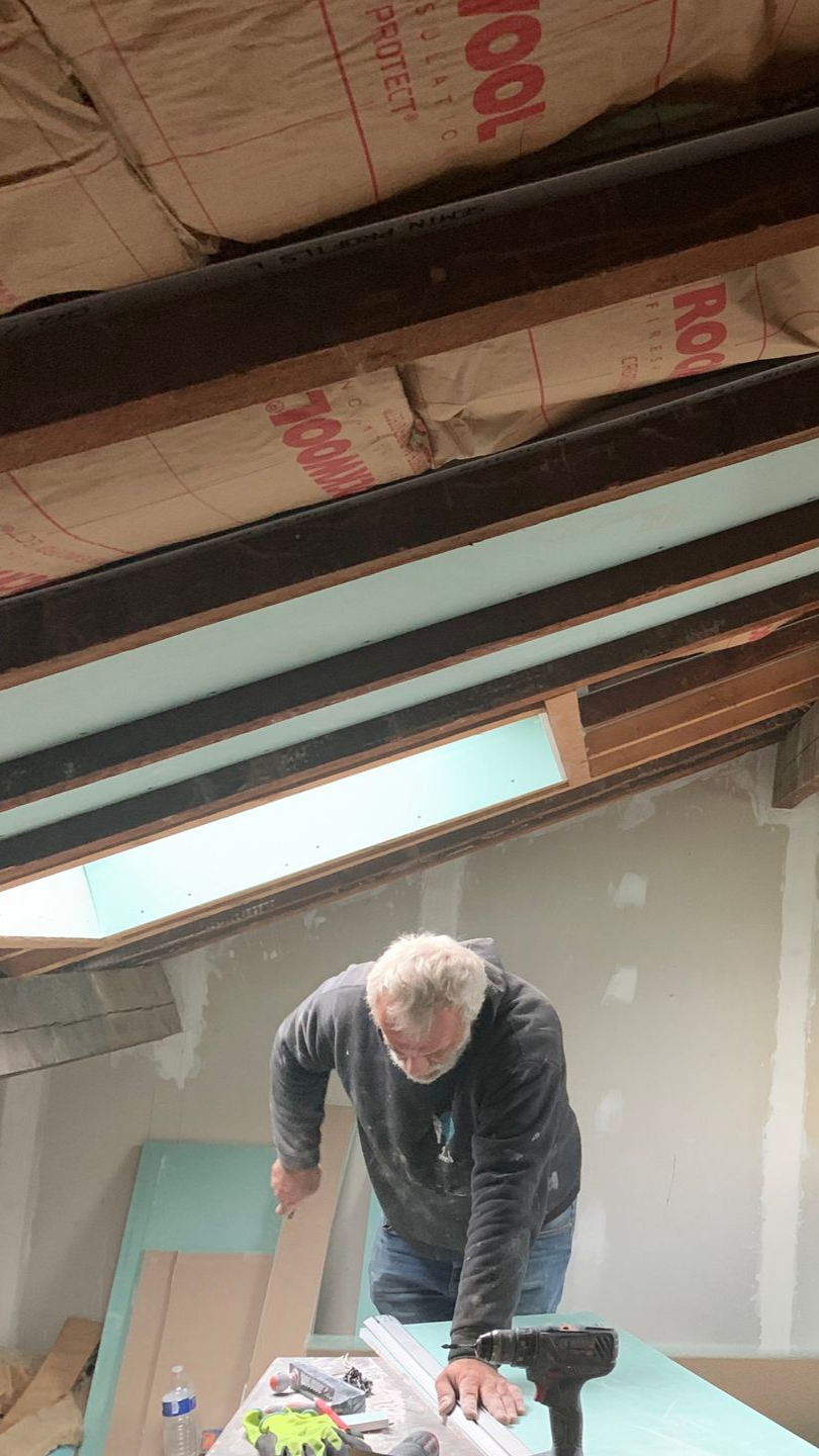 Un homme coupe du bois avec une perceuse dans une pièce aux poutres apparentes et à l'isolation défectueuse.