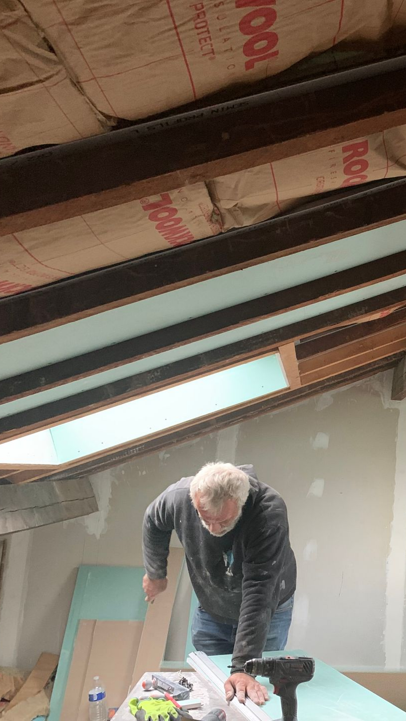Un homme travaille à l'isolation d'un grenier à l'aide d'une scie. Le grenier est doté d'un puits de lumière et de poutres apparentes.