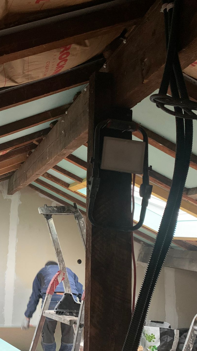 Une personne sur une échelle travaille sur un chantier de construction. On aperçoit des poutres en bois et des câbles électriques.