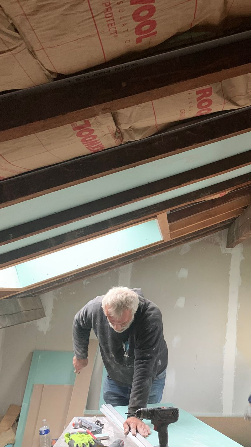 Un homme découpe de l'isolant en mousse dans un grenier avec des poutres apparentes et un puits de lumière.