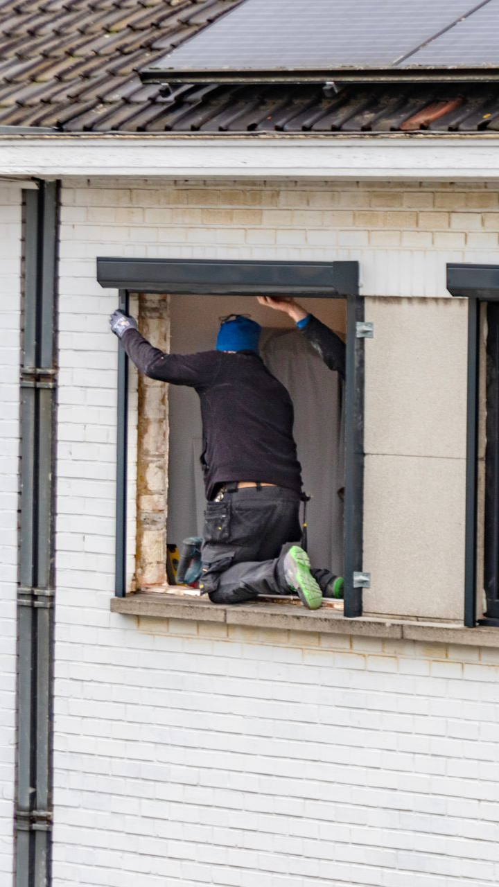 Une personne installe un volet. Elle est agenouillée dans l'encadrement de la fenêtre ouverte, vêtue de sombre et coiffée d'un chapeau bleu. Immeuble en briques.