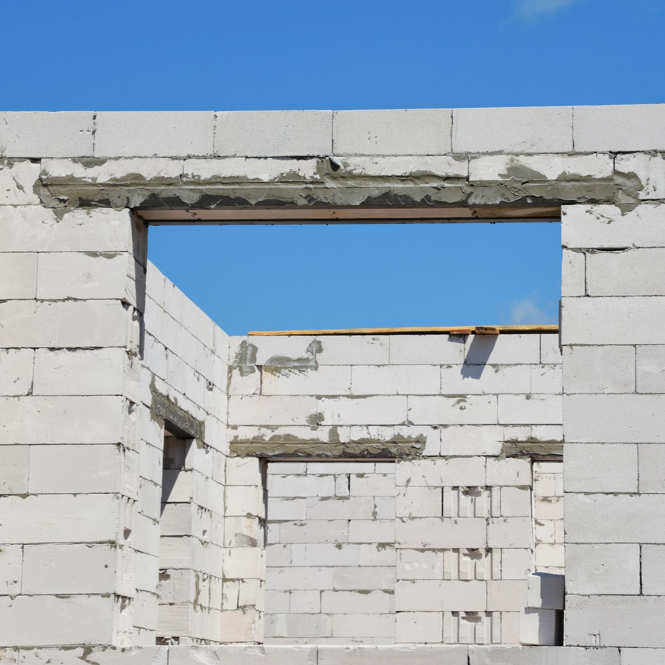 Intérieur d'un bâtiment inachevé, murs en parpaings de béton blanc, ouvertures rectangulaires pour les fenêtres, ciel bleu visible.