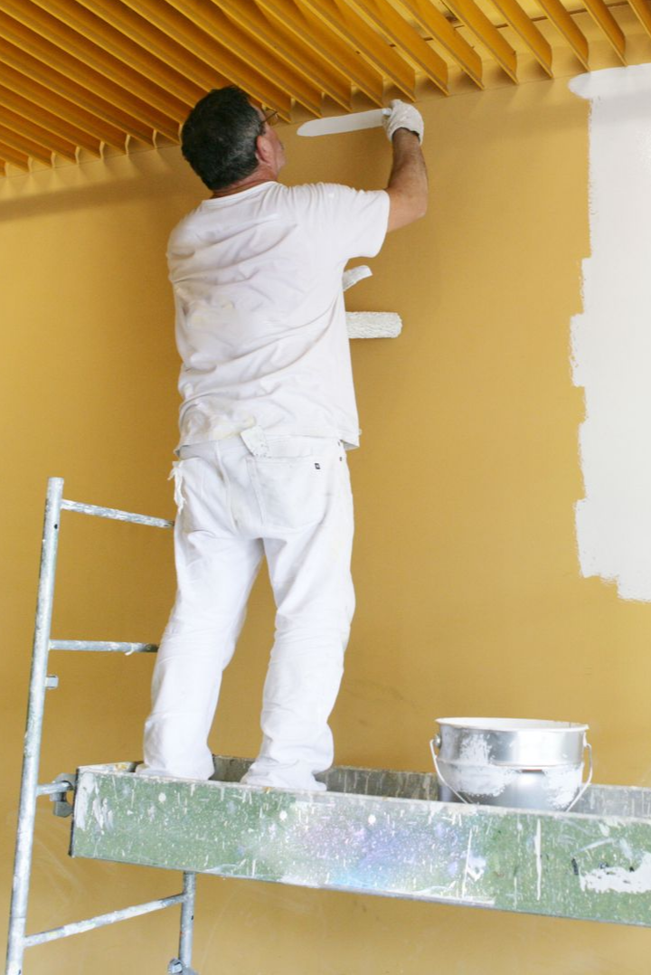 Peintre sur un échafaudage, peignant un mur et un plafond jaunes ; peinture blanche et vêtements.