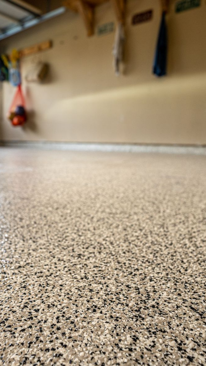 Gros plan sur un sol de garage beige et gris moucheté. Un mur flou et quelques objets suspendus apparaissent en arrière-plan.