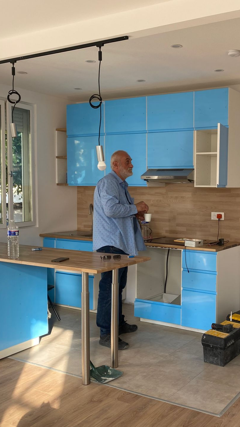 Un homme se tient debout dans une cuisine aux placards bleus. La cuisine est en travaux et on aperçoit des outils.