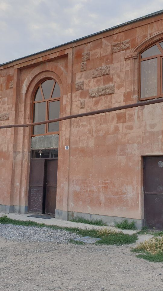 Bâtiment en pierre brun-rougeâtre avec une porte et une fenêtre cintrées. Un tuyau brun court le long de la façade. Espace vert devant.