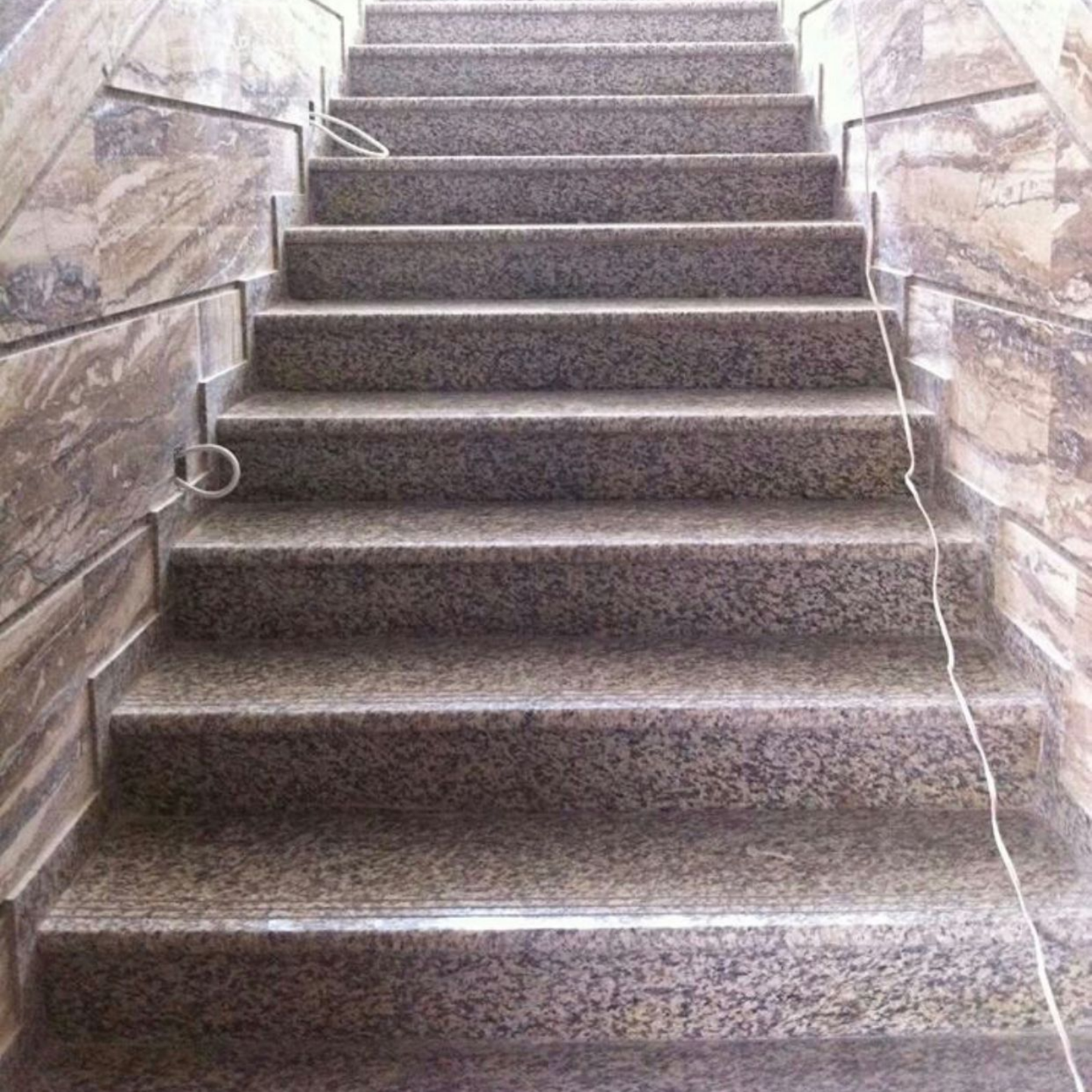 Escalier en pierre à motifs mouchetés bruns et gris, bordé de murs assortis. Fil électrique blanc visible.