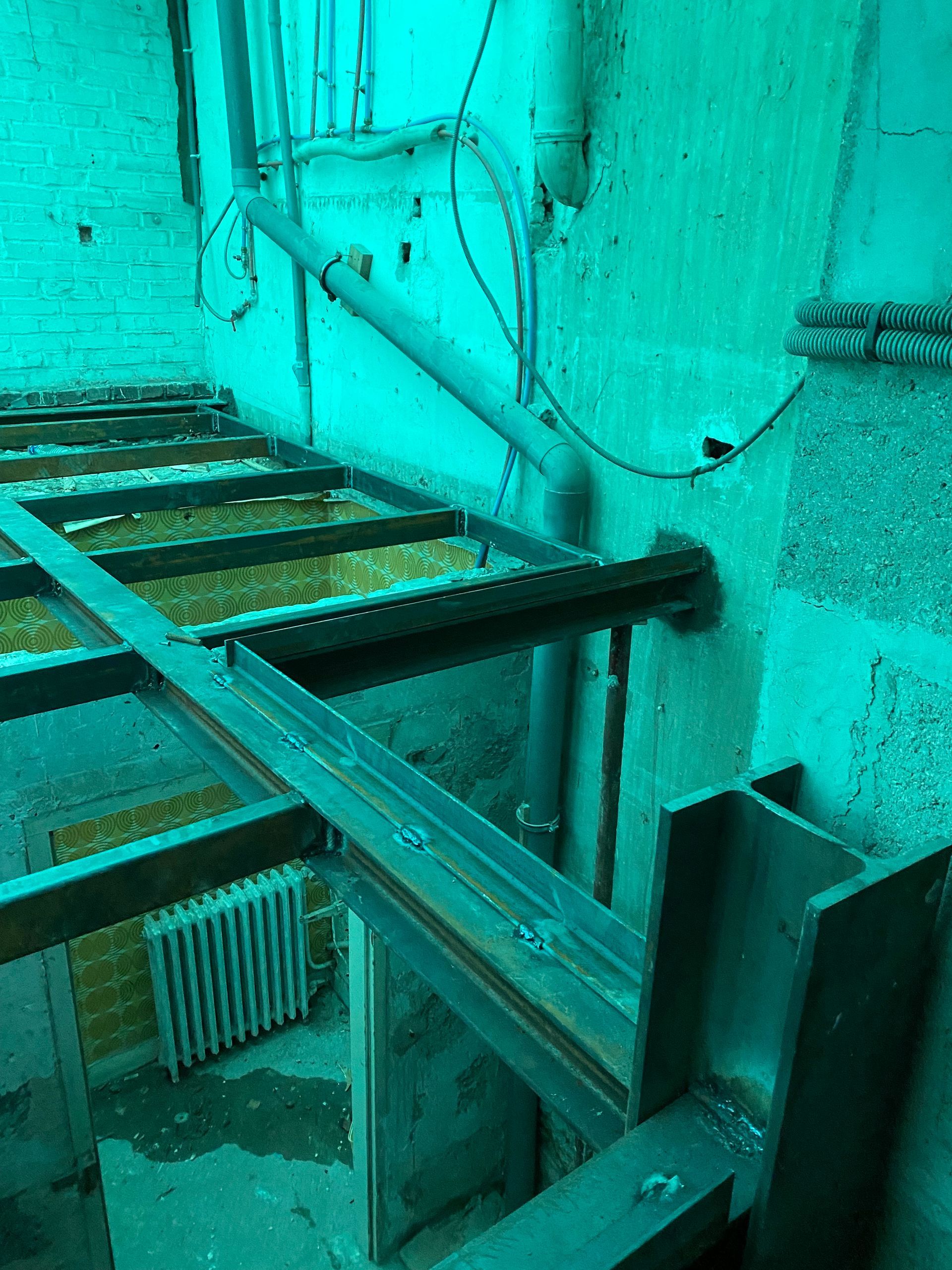 Une structure métallique avec des planches se trouve à l'intérieur d'une pièce où des tuyaux sont apparents au mur. Un radiateur est placé en dessous.