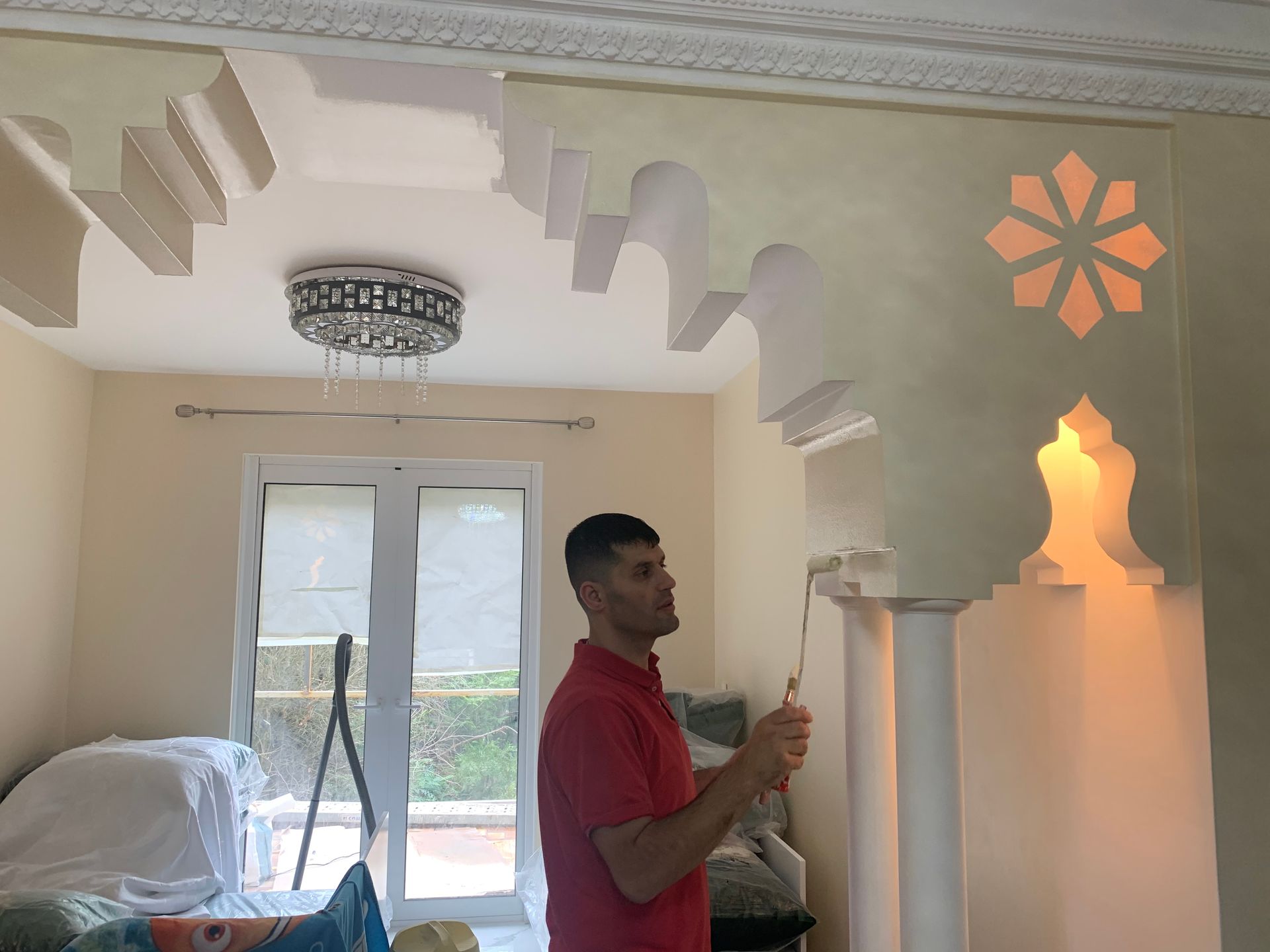 Un homme peint une arche architecturale ornée de découpes décoratives. Scène d'intérieur, éclairage chaleureux.