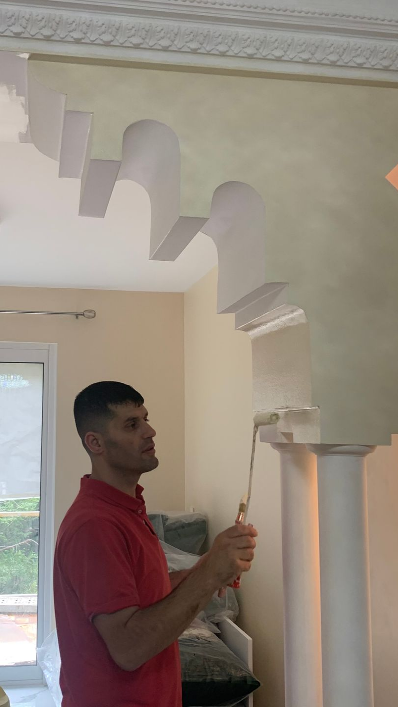 Un homme peint une arche décorative blanche au rouleau. Intérieur.