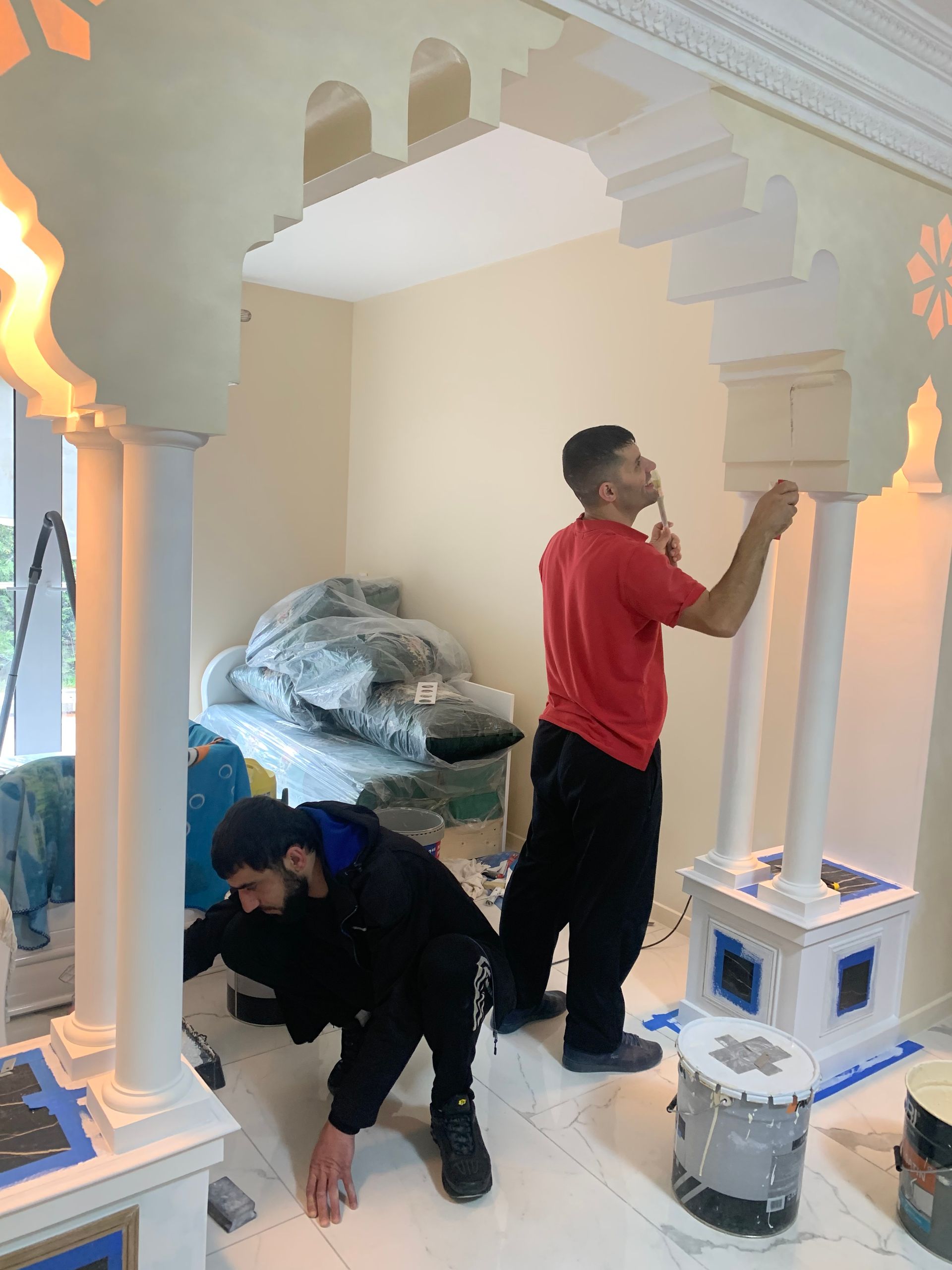 Deux ouvriers peignent l'encadrement d'une porte cintrée dans une pièce. L'un est debout, l'autre agenouillé. L'encadrement est blanc.