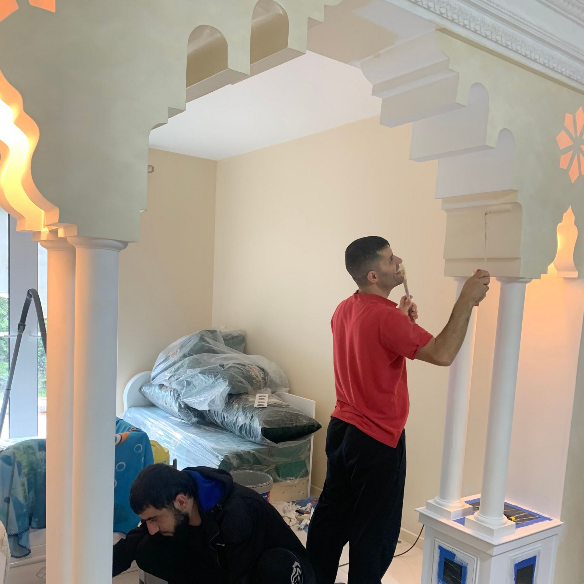 Deux personnes peignent une arche blanche ornée. L'une est accroupie, l'autre debout, toutes deux dans une pièce avec un lit.