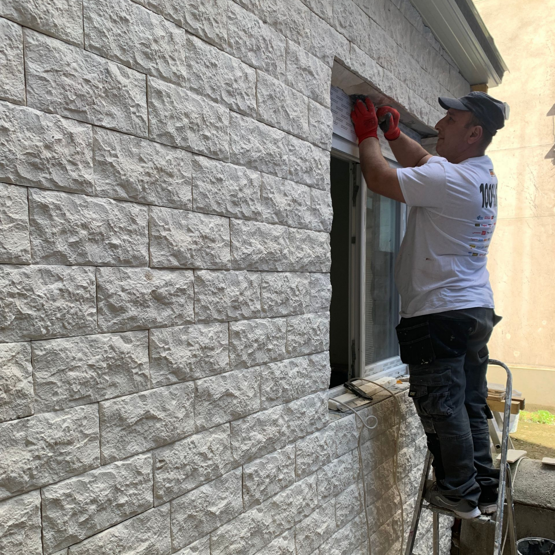Un homme installe des moulures autour d'une fenêtre sur un bâtiment à façade en pierre, portant des gants rouges et se tenant sur une échelle.