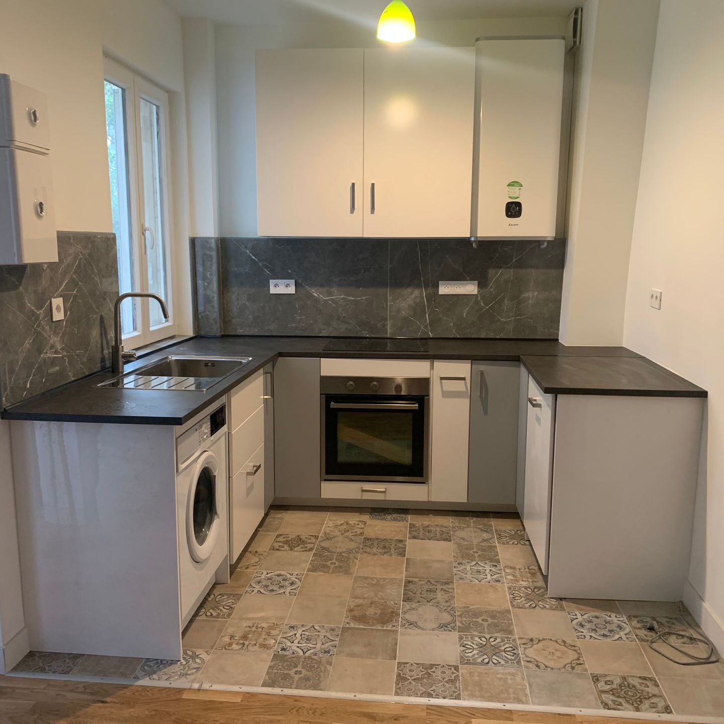 Petite cuisine avec placards blancs et gris, plans de travail foncés, sol carrelé et électroménager.