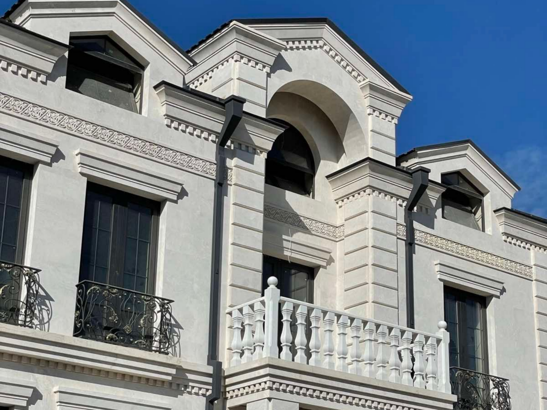 Bâtiment blanc aux ornements travaillés, avec une fenêtre cintrée et un petit balcon aux balustrades en fer forgé noir.