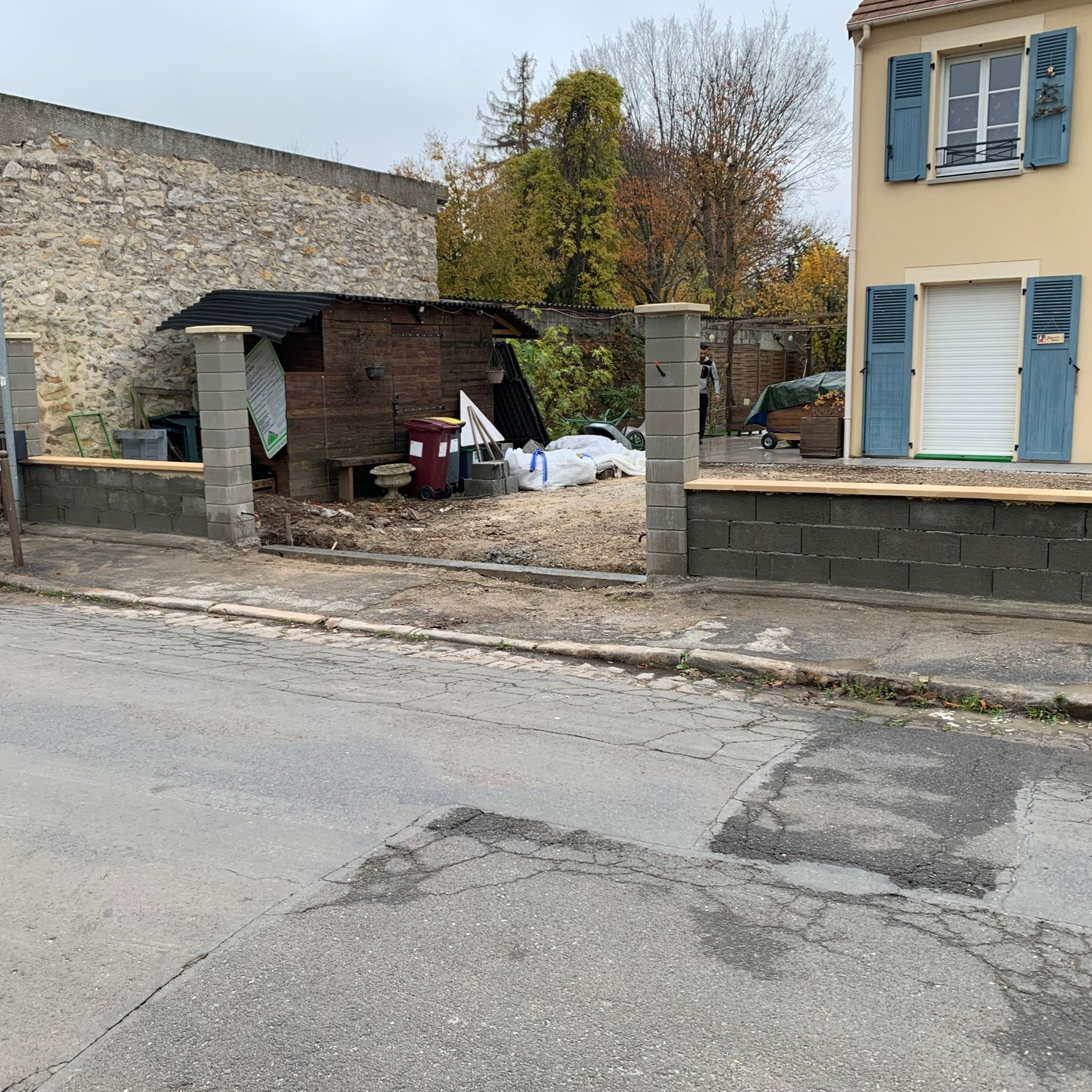 Vue de rue avec un mur de parpaings, un abri de jardin et une maison beige aux volets bleus. Journée nuageuse.