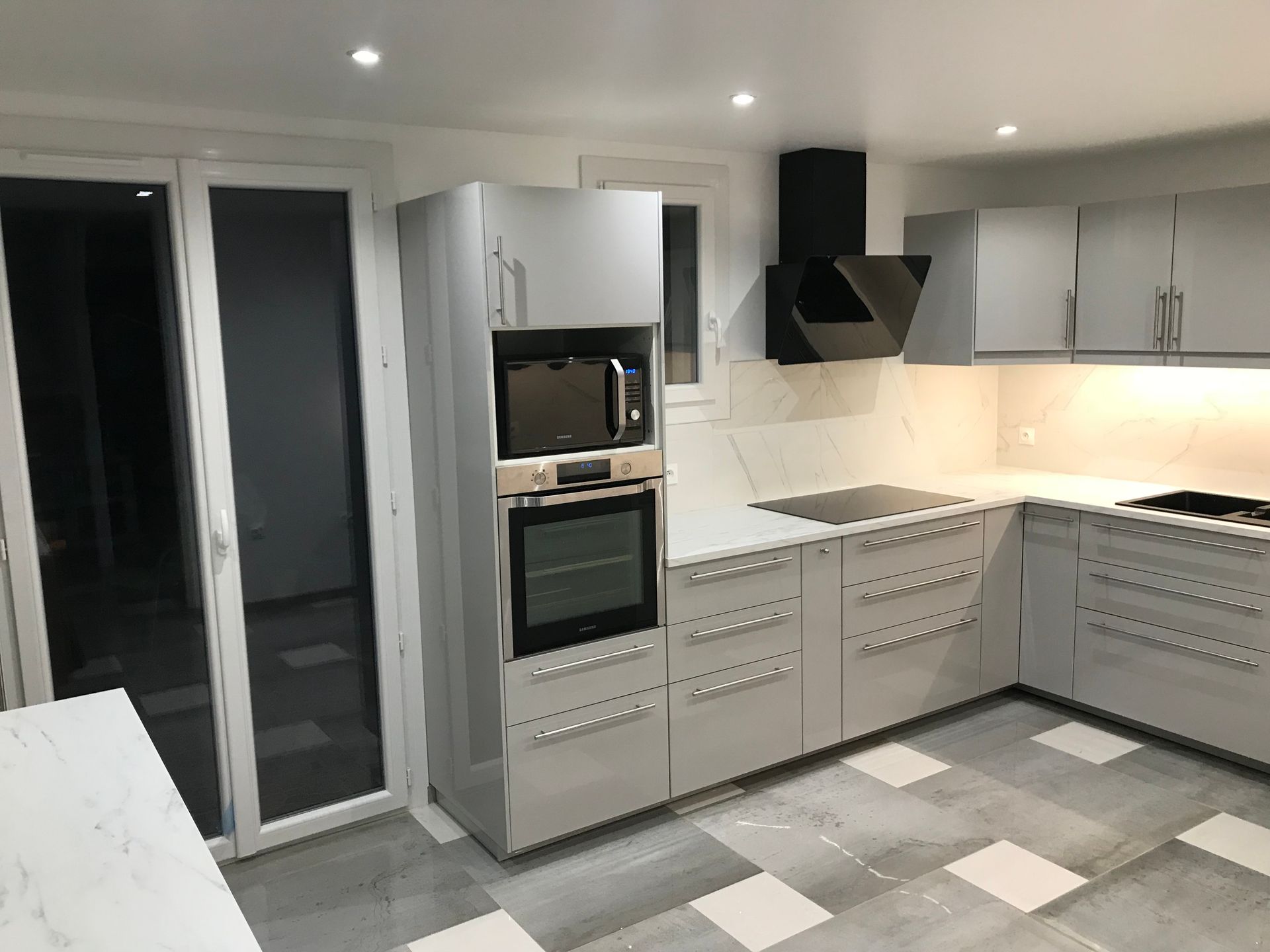 Cuisine moderne grise avec appareils électroménagers intégrés, comptoirs blancs et sol carrelé, à côté d'une porte vitrée.