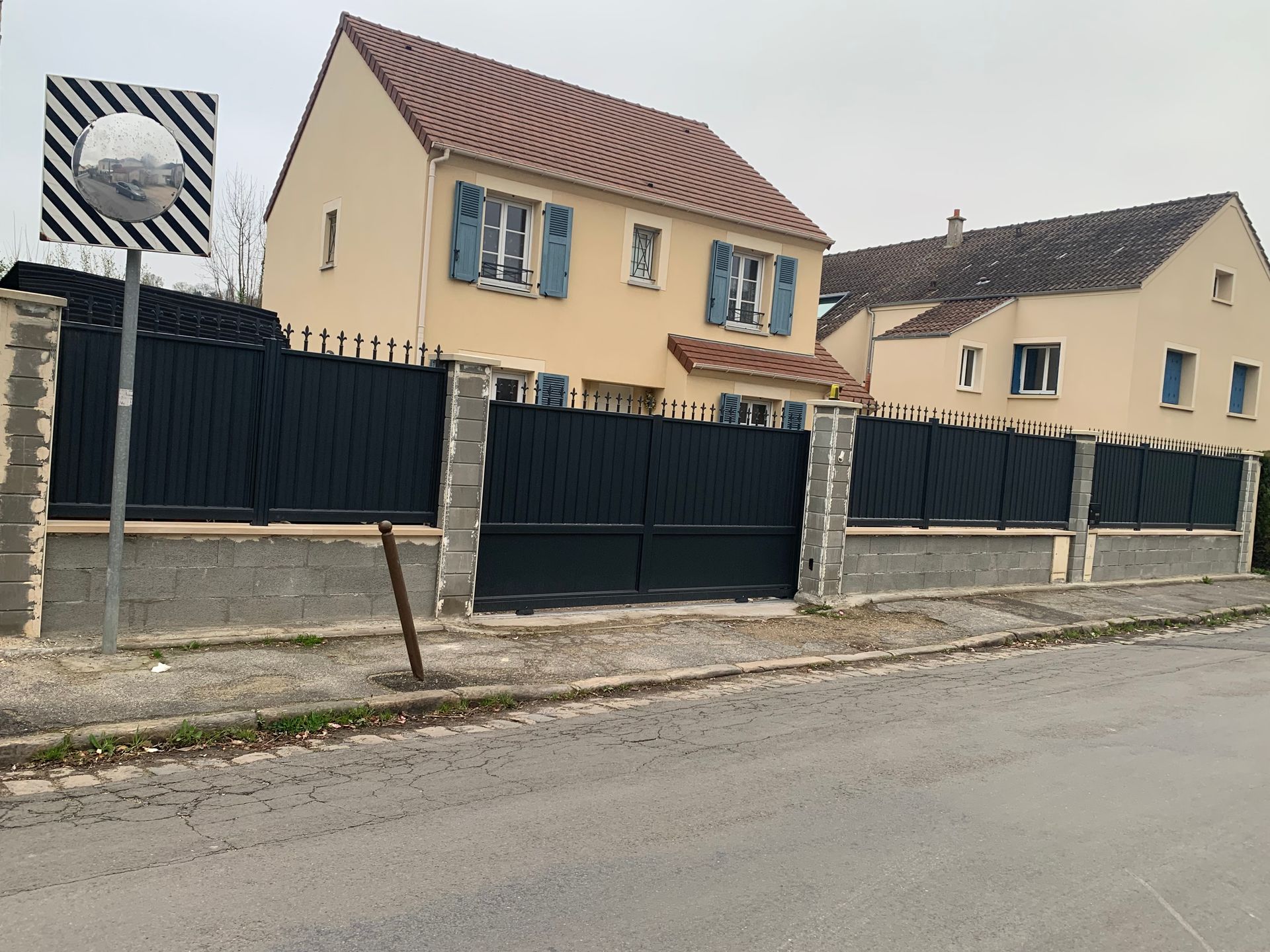 Une maison beige à deux étages derrière une clôture noire, dans une rue grise sous un ciel nuageux.
