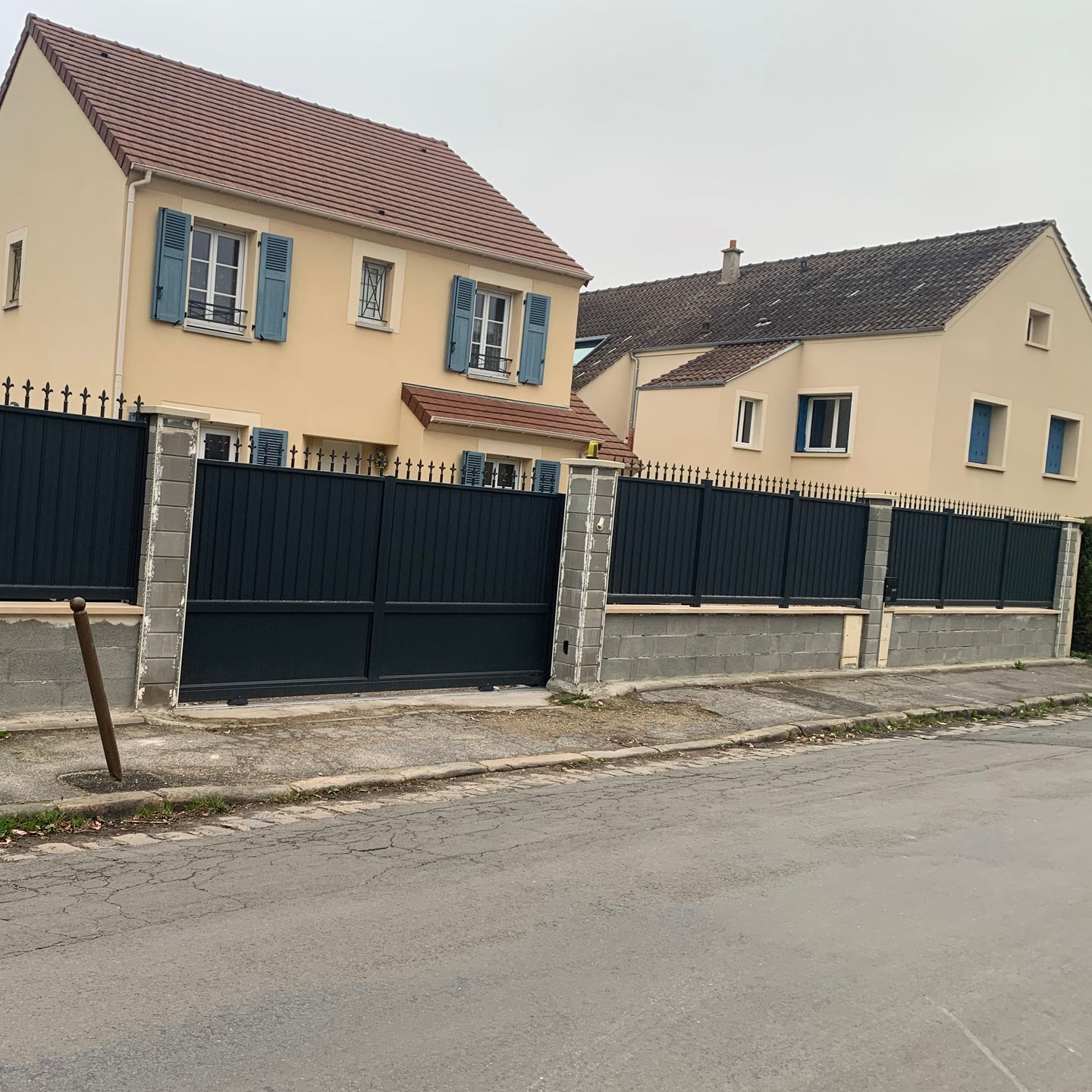 Des maisons à deux étages se dressent derrière une clôture sombre aux piliers de béton, dans une rue au ciel couvert.