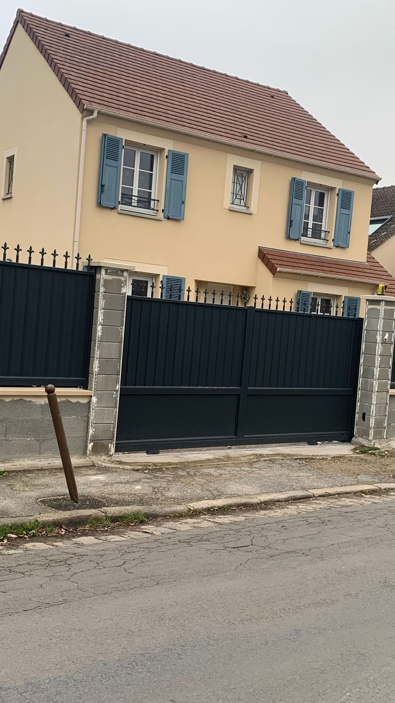 Maison beige à deux étages avec des volets bleus, un portail sombre et des poteaux en béton.