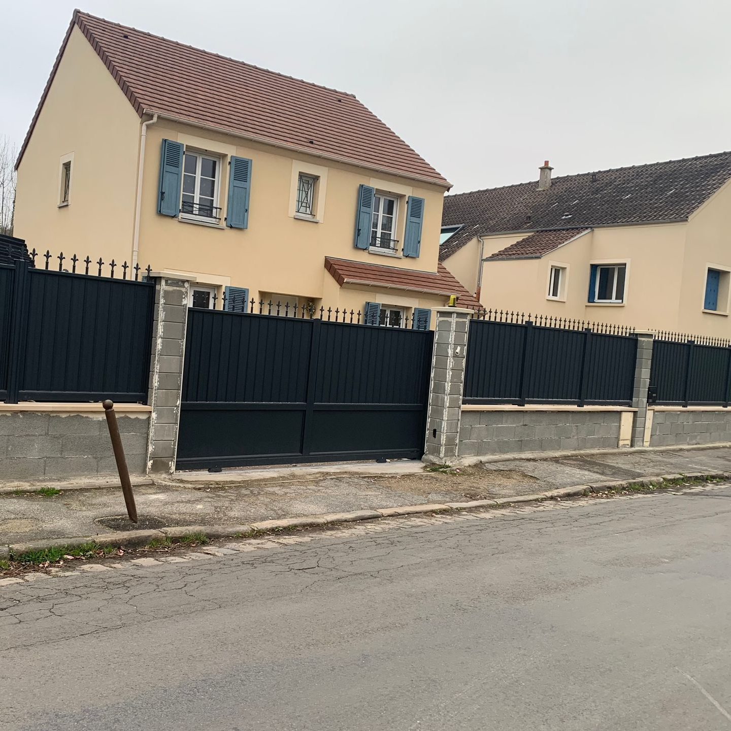 Maisons beiges aux volets bleus derrière une clôture gris foncé aux piliers en béton.