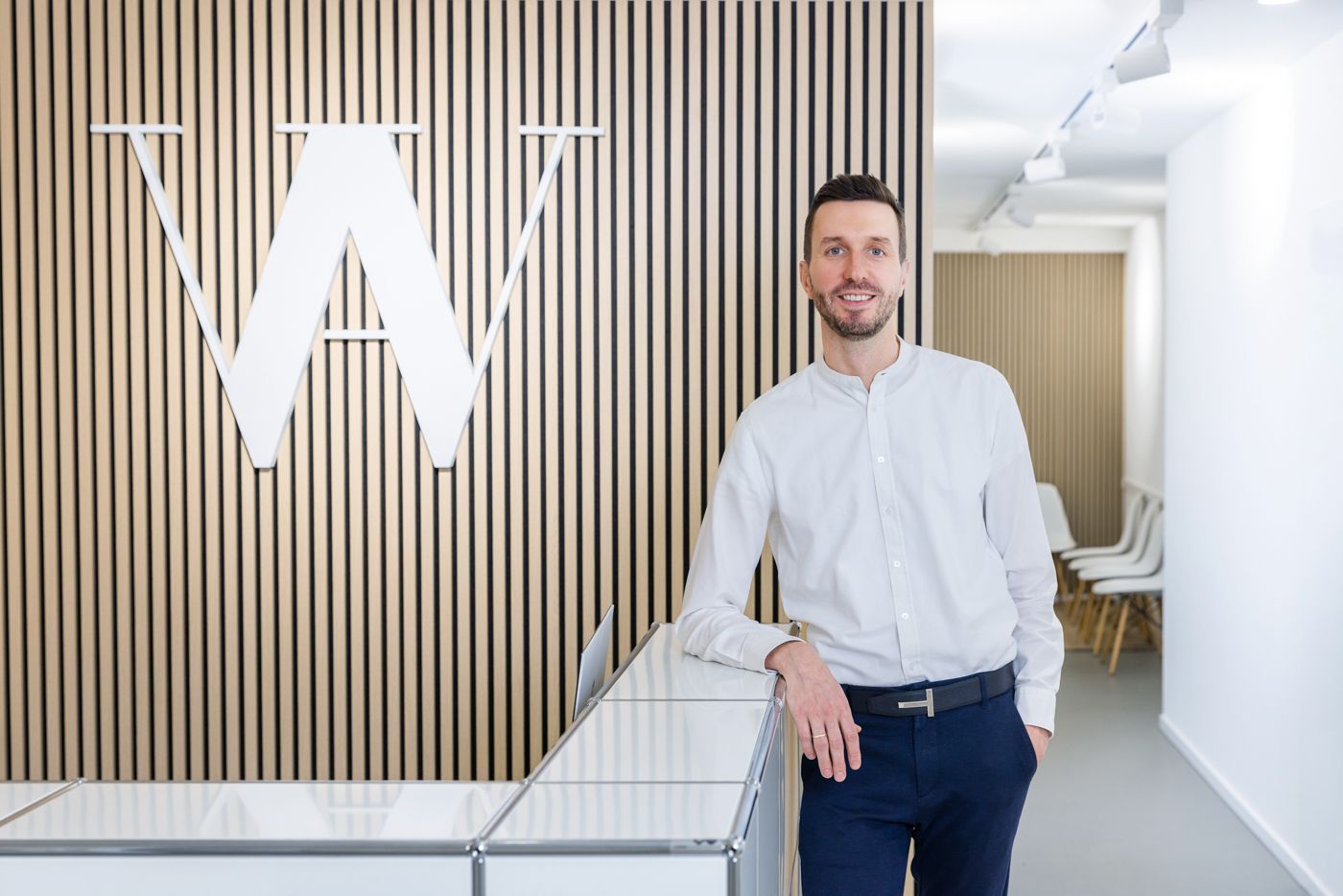 Mann im weißen Hemd lächelt neben einem Rezeptionstresen mit einem Logo. Holzpaneelwand.
