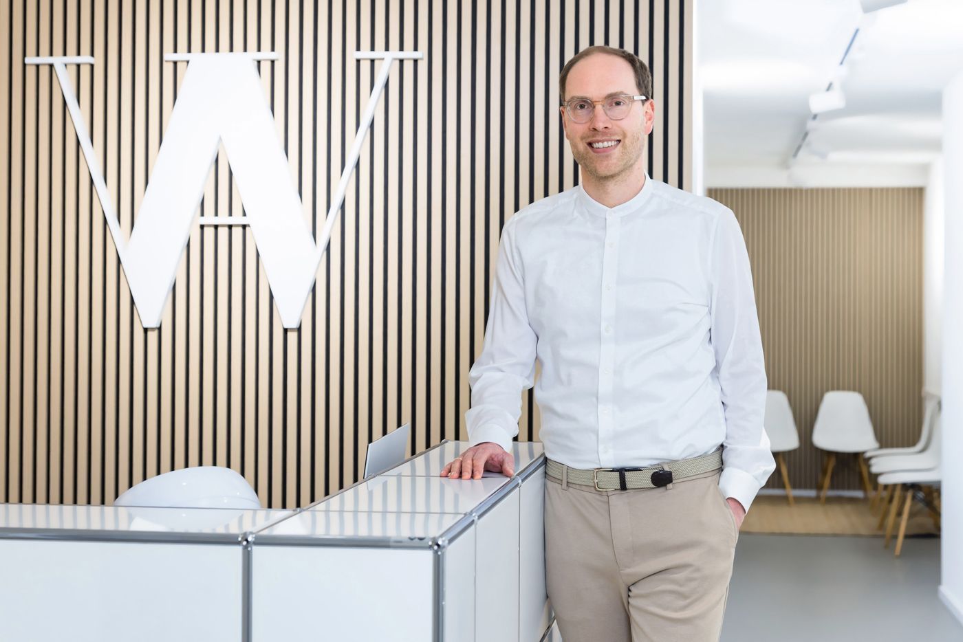 Lächelnder Mann mit Brille steht an der Rezeption. Weißes Hemd, beige Hose. Holzwand mit Logo.