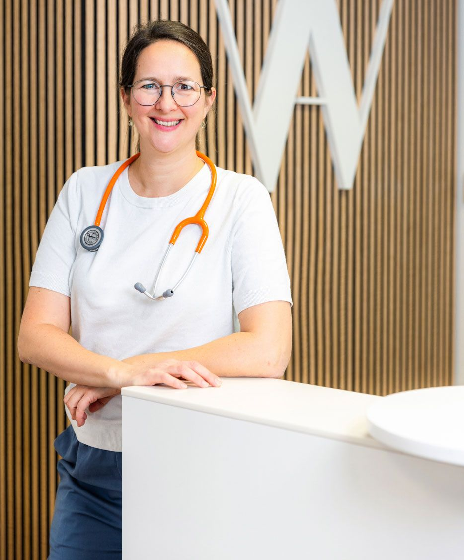 Frau an der Rezeption lächelt, weißes Oberteil, holzgetäfelte Wand mit „W“-Logo, Stühle sichtbar.