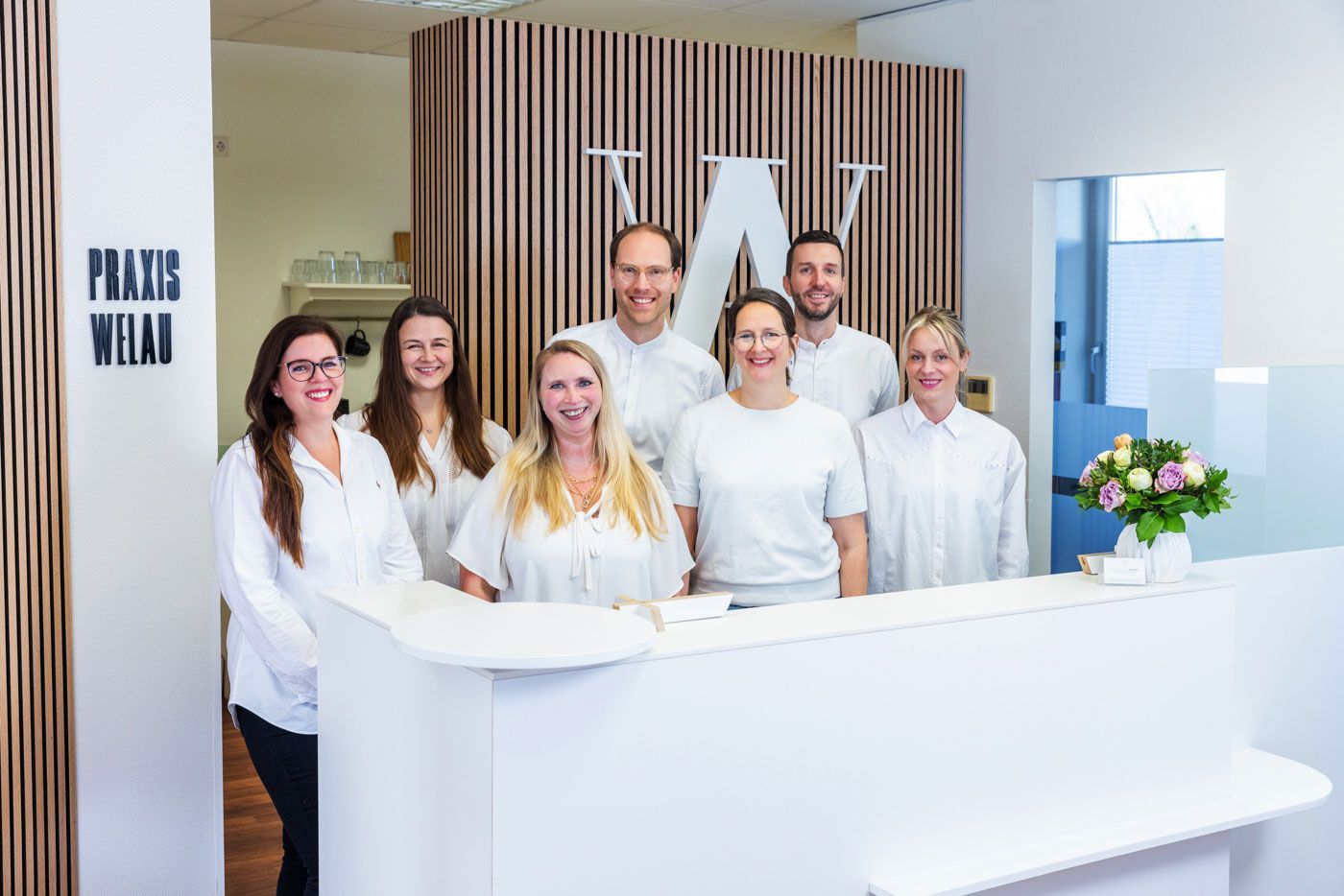 Gruppe von Menschen in weißen Hemden hinter einem weißen Empfangstresen mit einem Logo in einem hellen Büro.