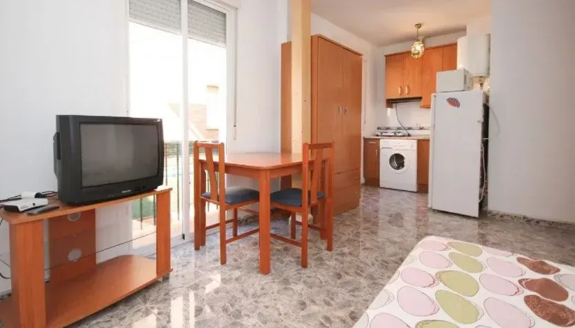 Pequeño apartamento tipo estudio con TV, mesa con sillas y cocina americana.