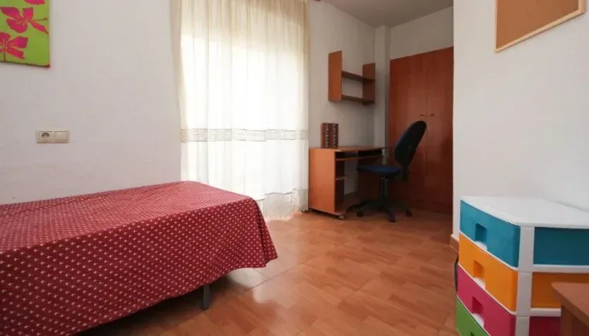 Dormitorio con colcha roja, escritorio y cajones de colores, junto a una ventana con cortinas.
