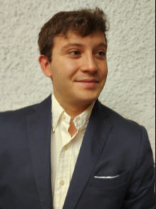 Un hombre con un blazer azul y una camisa abotonada de color amarillo claro sonríe, mirando ligeramente hacia un lado.