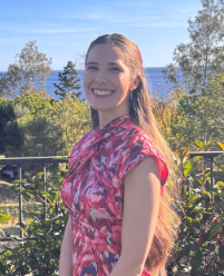 Mujer sonriendo, vistiendo un vestido estampado de color rosa, de pie al aire libre con árboles y agua en el fondo.