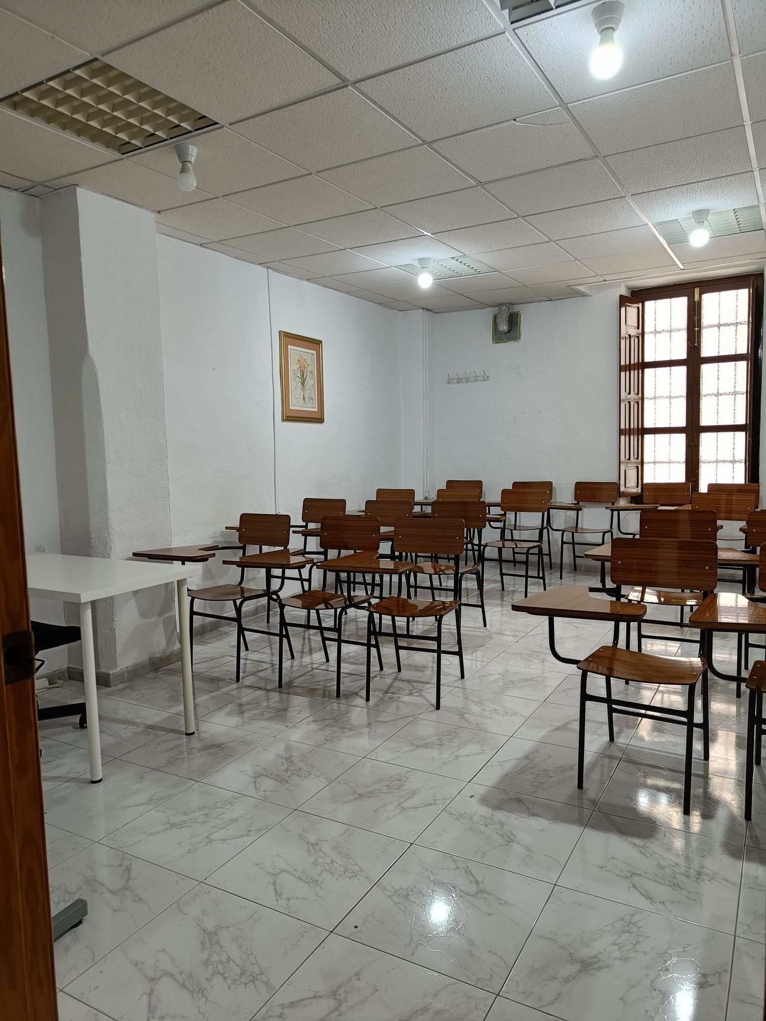 Aula vacía con filas de pupitres y una mesa blanca. Madera marrón claro, paredes blancas y suelo de baldosas.