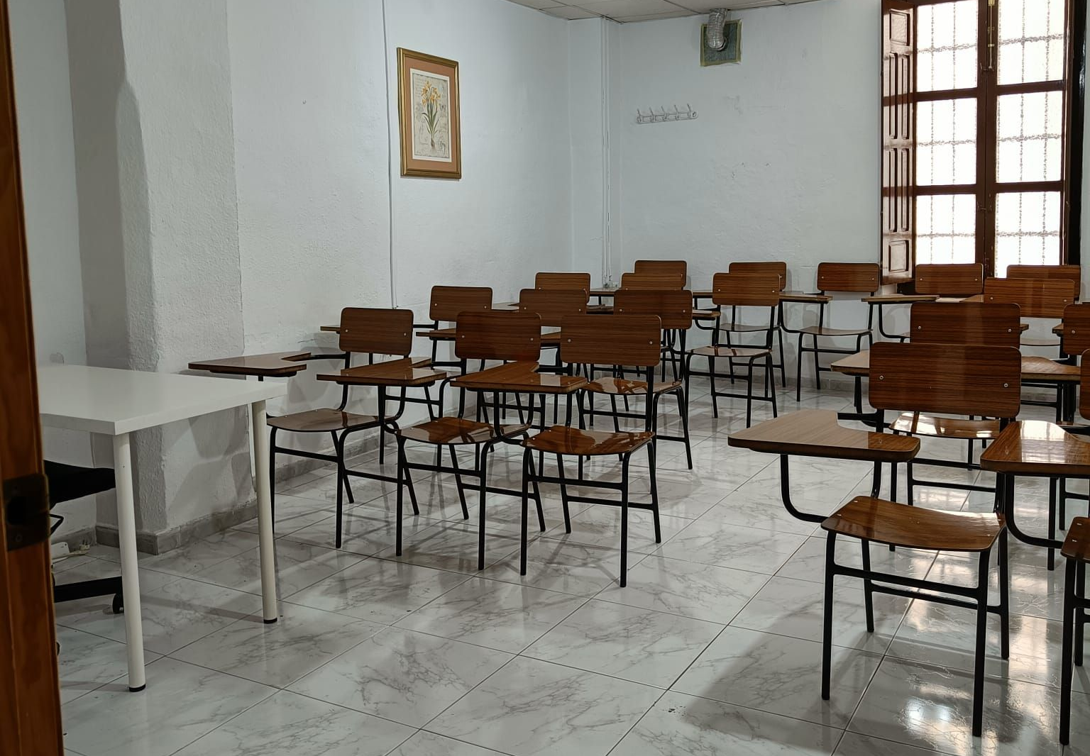 Aula vacía con filas de pupitres y una mesa blanca. Madera marrón claro, paredes blancas y suelo de baldosas.