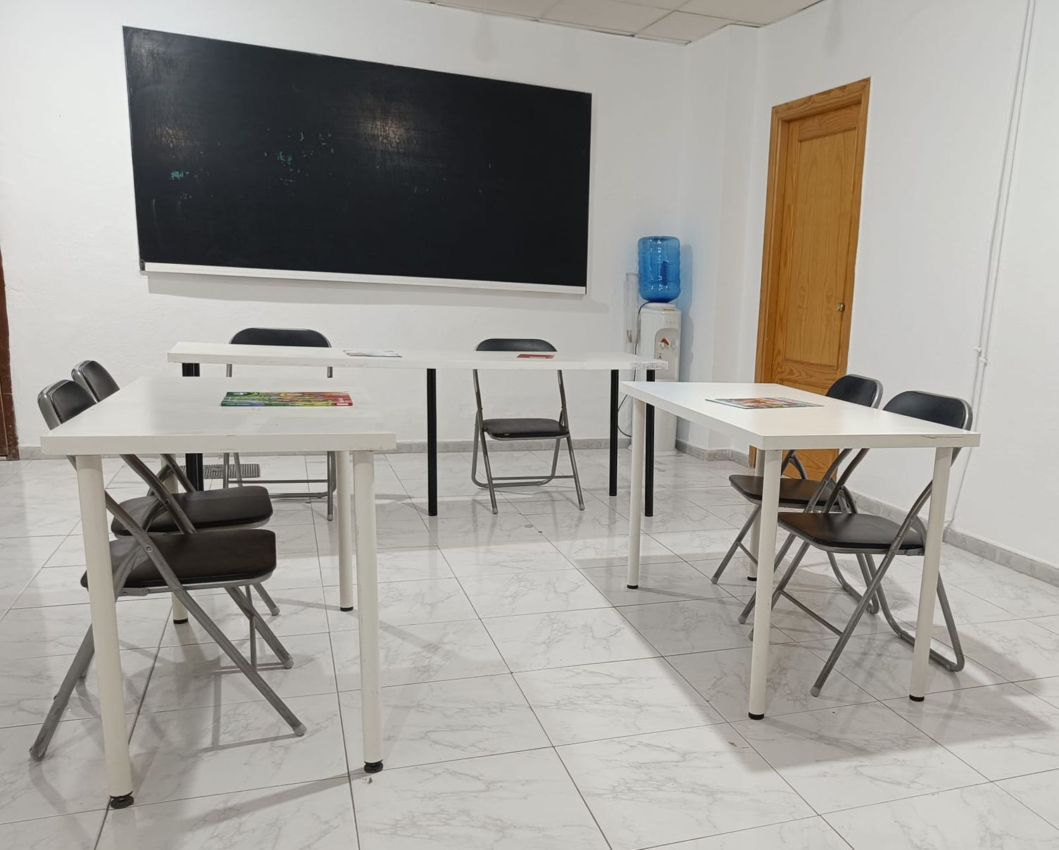 Aula vacía con mesas, sillas y pizarra. Paredes blancas, suelo de baldosas y dispensador de agua.