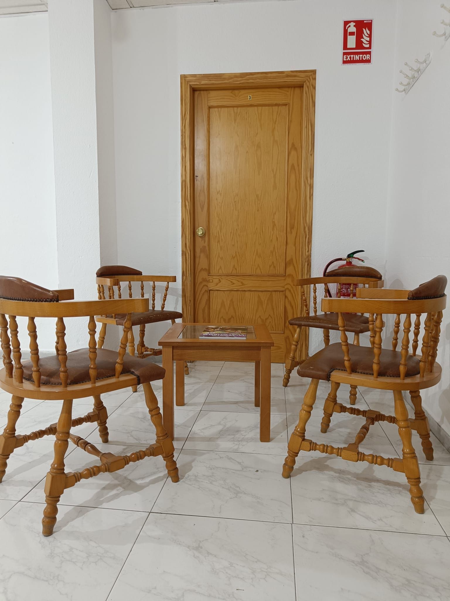 Una pequeña mesa de madera y cuatro sillas lo rodean. Al fondo, una puerta cerrada.